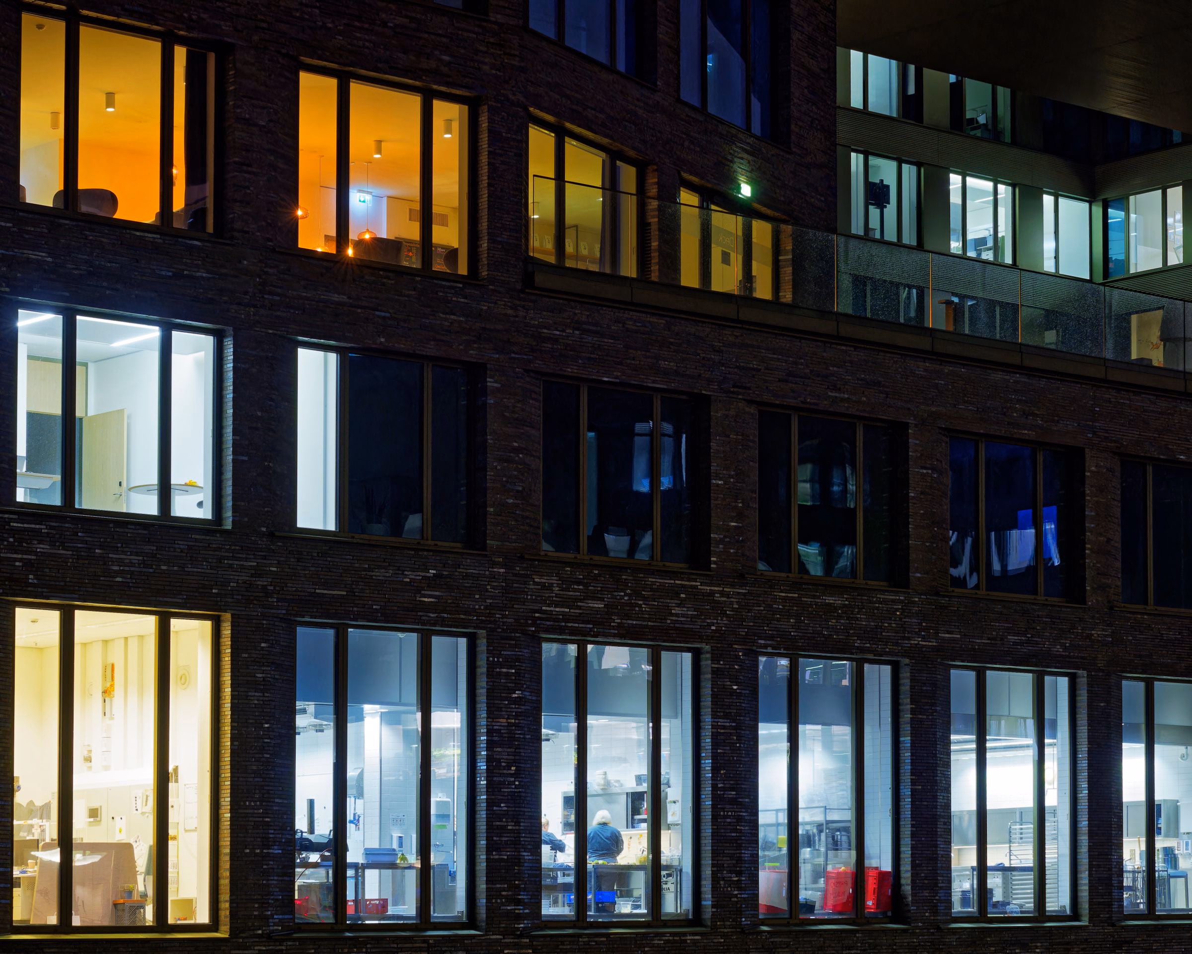 Glass-and-steel commercial building at dusk, every floor lit, viewed from the street level looking up.